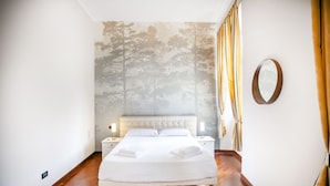A bedroom with a large bed, wooden flooring, a nightstand, a wall-mounted mirror, and a window with curtains.
