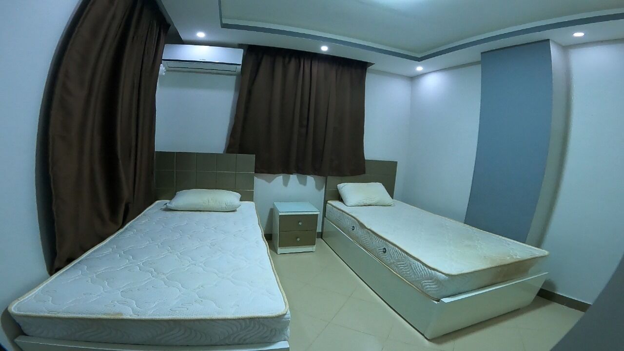 A hotel room with two single beds, a small bedside table, a wall-mounted air conditioner, and brown curtains.