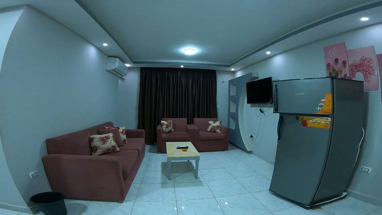 A living room with a sofa set, a coffee table, a refrigerator, and a TV mounted on the wall.