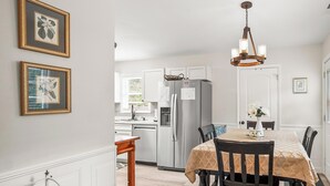 A modern kitchen with white cabinetry, a stainless steel refrigerator, and a dining area with a table set for four. - Cozy Cottage, Deck + Yard, Near Downtown & 10 min to Parris Island (Beaufort)