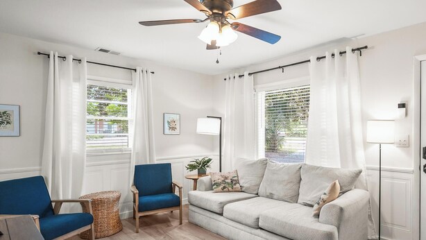 A modern living room with a ceiling fan, a blue armchair, a grey sofa, and a small side table with a plant. - Cozy Cottage, Deck + Yard, Near Downtown & 10 min to Parris Island (Beaufort)