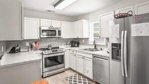 A modern kitchen with white cabinets, stainless steel appliances, and a wooden dining table. - Cozy Cottage, Deck + Yard, Near Downtown & 10 min to Parris Island (Beaufort)