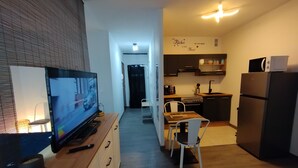 Ein kompakter Wohnbereich mit Fernseher auf einer Holzkommode, einem Essbereich mit Tisch und Stühlen, einer Küche mit modernen Geräten und einem Flur, der in einen weiteren Raum führt.