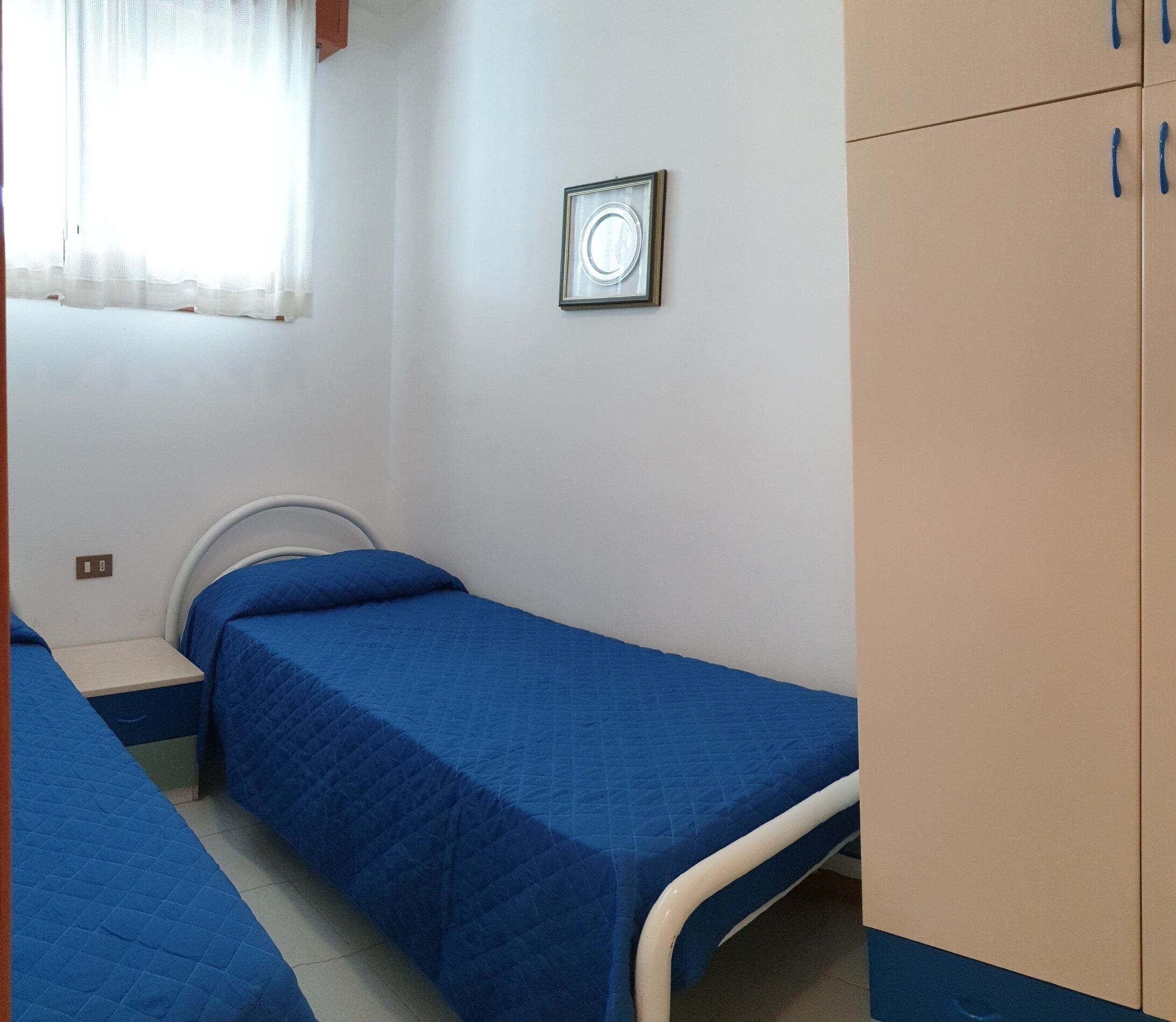Una habitación pequeña con cama individual, colcha azul, cabecero blanco y armario de madera.