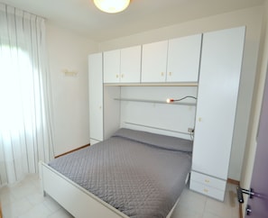 A bedroom with a bed, white wardrobe, and a window with curtains. - Coastal Escape Flat With Common Pool (San Michele al Tagliamento)