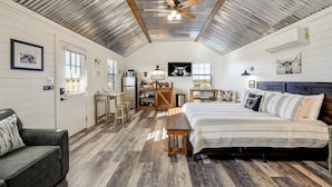 A spacious room with a high ceiling, a bed, a sofa, a desk, and a kitchen area. - Windmill Meadows Cottages-firepit-hill Country Views!! (Harper)