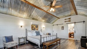 A spacious room with a high ceiling, a bed, a bench, and a chair. - Wine Country Cottage With Scenic Hill Country Views (Harper)