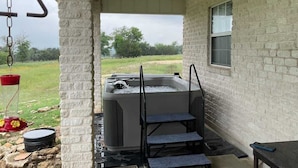 A hot tub with steps, a window, and a hanging light fixture in a covered outdoor area with a brick wall.