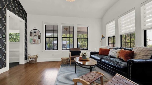 A spacious living room with a black leather sofa, a wooden bench, and a coffee table. There are three large windows with black frames and a chandelier hanging from the ceiling.