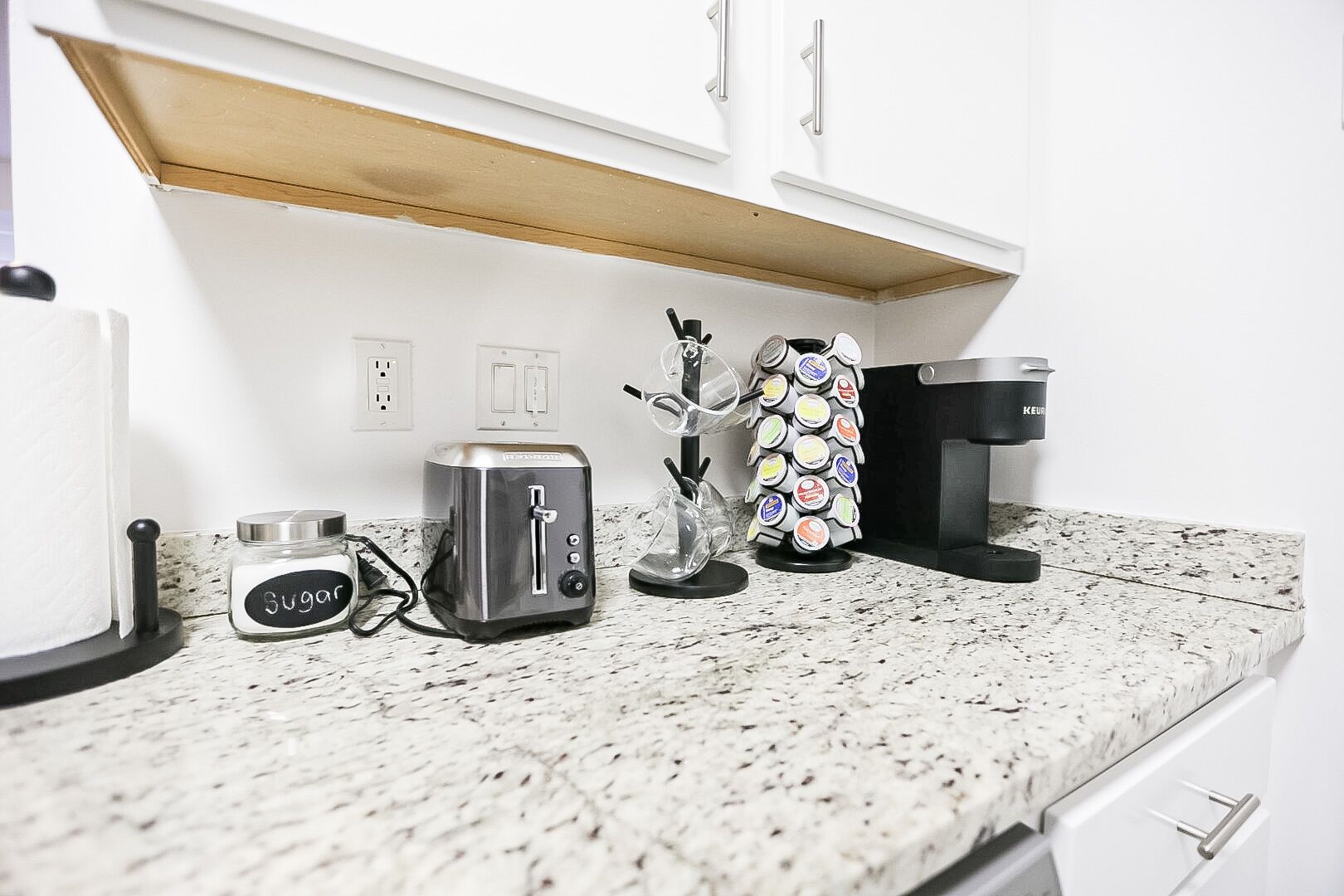 Una cocina con tostadora, cafetera, hervidor de agua y un tarro etiquetado como 'Sugar'.