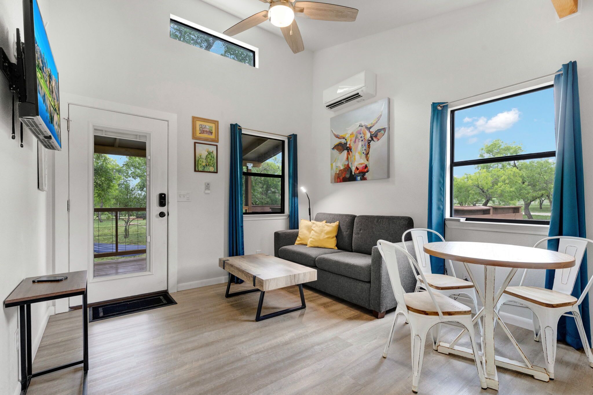 A modern living room with a sofa, a coffee table, and a dining area with chairs. There is a ceiling fan, a window with curtains, and artwork on the walls.