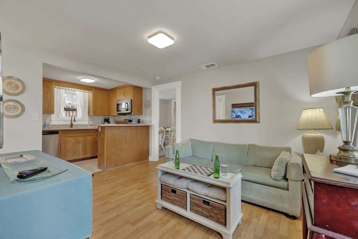 A living room with a sofa, a coffee table, and a TV. There is a kitchen in the background with wooden cabinets and a microwave.