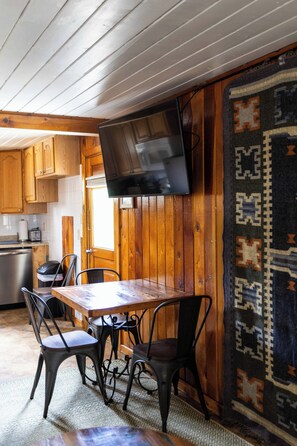 A wooden interior with a dining table, chairs, a television mounted on the wall, and a patterned rug.