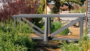 Eine Holzbrücke über einen kleinen Bach, umgeben von viel Grün, mit einem Haus im Hintergrund.
