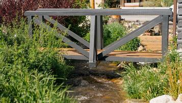 Un pont en bois enjambant un petit ruisseau, entouré de verdure, avec une maison en arrière-plan.