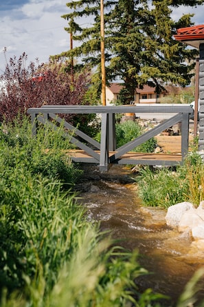 Un puente de madera cruzando un arroyo, con vegetación a su alrededor y una casa al fondo.