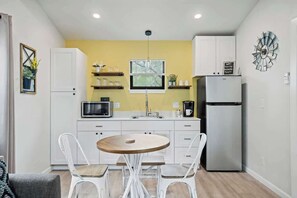 A compact kitchen with white cabinets, a microwave, a refrigerator, and a dining table with chairs. - Winecup Waterfront Cottage (Fredericksburg)