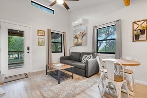 Una sala moderna con un sofá gris, una mesa de centro de madera, una mesa de comedor blanca con sillas y ventanas amplias con vistas a árboles.
