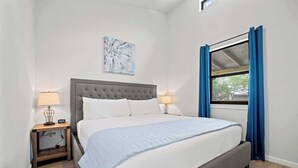 A bedroom with a large bed, two bedside tables, a window with curtains, and a wall-mounted artwork. - Winecup Waterfront Cottage (Fredericksburg)