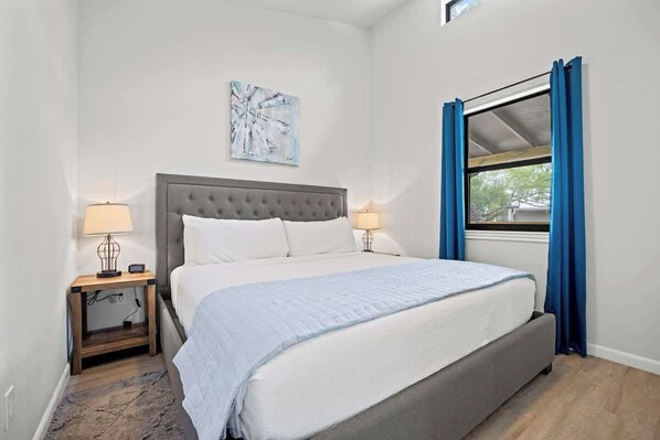 A bedroom with a large bed, two bedside tables, a window with curtains, and a wall-mounted artwork. - Winecup Waterfront Cottage (Fredericksburg)