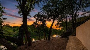 A wooden bench, trees, and a view of a body of water during sunset. - Rivers Edge Cottages With Hot Tub (Fredericksburg)