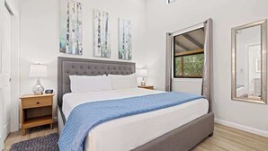 A bedroom with a bed, two bedside tables, a window with curtains, and wall art. - Rivers Edge Cottages With Hot Tub (Fredericksburg)