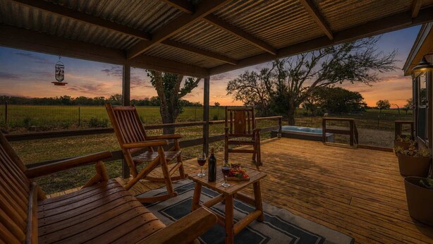 A covered outdoor seating area with wooden furniture, a hot tub, and a view of a field at sunset. - Rivers Edge Cottages With Hot Tub (Fredericksburg)