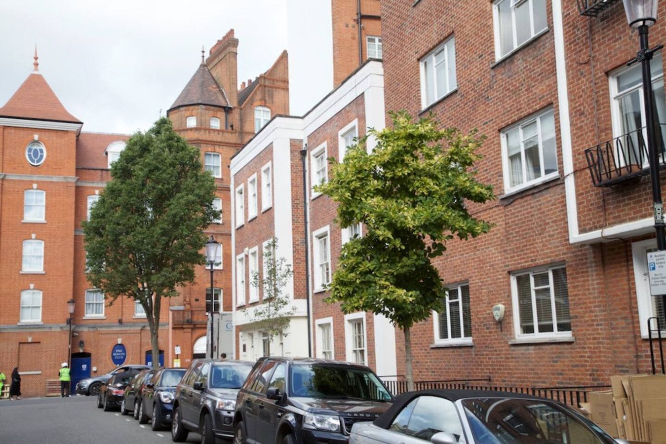 Une rangée de voitures garées le long d’une rue bordée d’immeubles en briques, dont un bâtiment doté d’une tour et d’une enseigne sur laquelle on peut lire « The Club ».