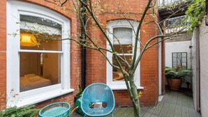 A narrow courtyard with a brick building, a tree, and two blue chairs. - Stunning 3 bed flat in the heart of Chelsea (London)