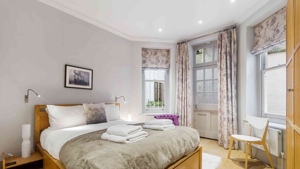 A bedroom with a large bed, a bedside table, a chair, a window with curtains, and a radiator. - Stunning 3 bed flat in the heart of Chelsea (London)