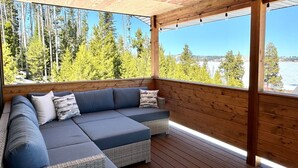 A covered outdoor seating area with a sectional sofa, wooden deck, and a view of a forest and water. - Fall Dates Available for Epic Sunsets (Anaconda)