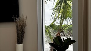 A room with a wooden table, a white vase with dried plants, a potted plant with white pebbles, and a view of palm trees and a body of water through a window. - Elegante Casa con Vistas al mar en la Playa #7 (Urbanova)
