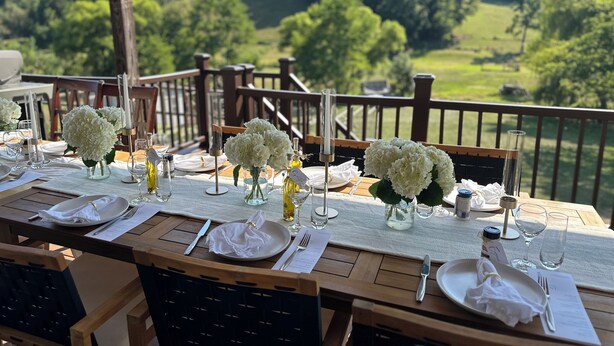 Un área de comedor al aire libre con mesas dispuestas para una comida, rodeada por una terraza de madera y una vista panorámica de un paisaje verde.