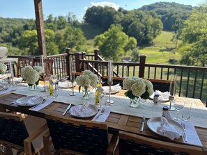 An outdoor dining area with tables set for a meal, surrounded by a wooden deck and a scenic view of a green landscape.