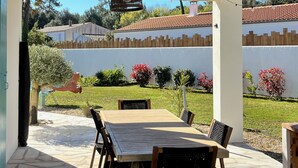 Une terrasse extérieure avec une table et des chaises en bois, un luminaire suspendu et une vue sur un bâtiment blanc avec un toit recouvert de tuiles rouges en arrière-plan.