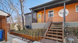 A two-story building with a wooden front door, a staircase with a metal railing, and a sign that reads "The Mural." - Mural House A: On The Trails > Private Hot Tub > Fire Pit > EV Charger (Bentonville)