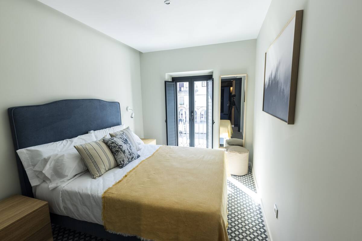 Ein Schlafzimmer mit einem Bett, Nachttischen, einem Fenster und einer Tür zu einem anderen Raum.