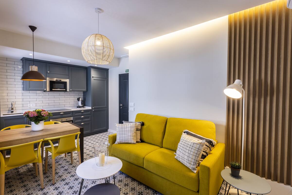 Ein modernes Wohnzimmer mit einem gelben Sofa, einem Esstisch mit gelben Stühlen, einer Küchenzeile mit grauen Schränken und einem gemusterten Boden.