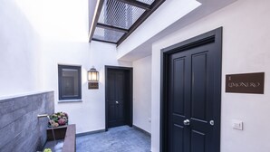 Un couloir étroit avec deux portes noires, un banc, un bac à fleurs et un luminaire fixé au mur.