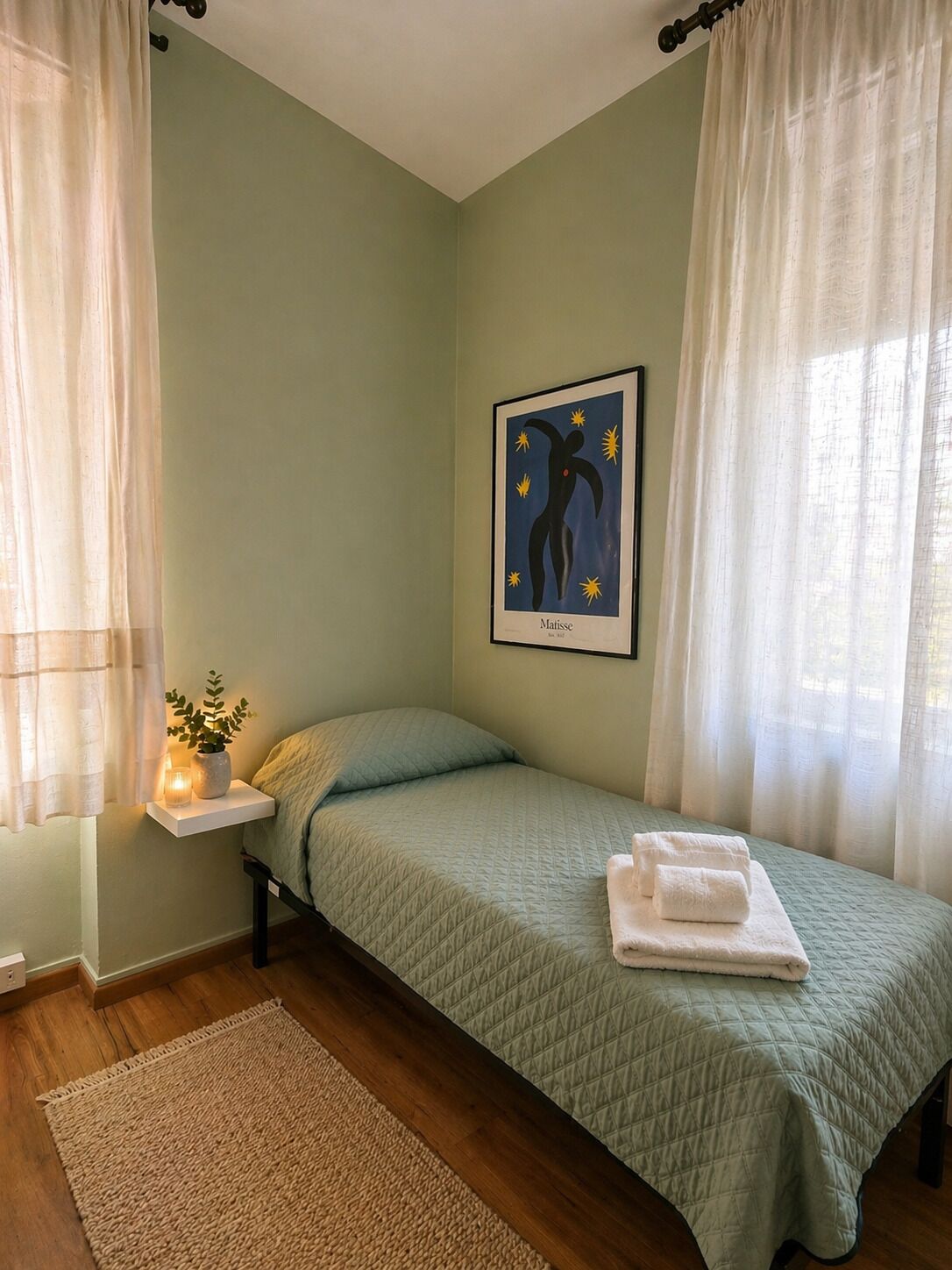 A single bed with a quilted green bedspread, a bedside table with a candle, and a framed abstract art piece on the wall.