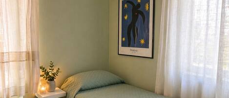 A single bed with a quilted green bedspread, a bedside table with a candle, and a framed abstract art piece on the wall.