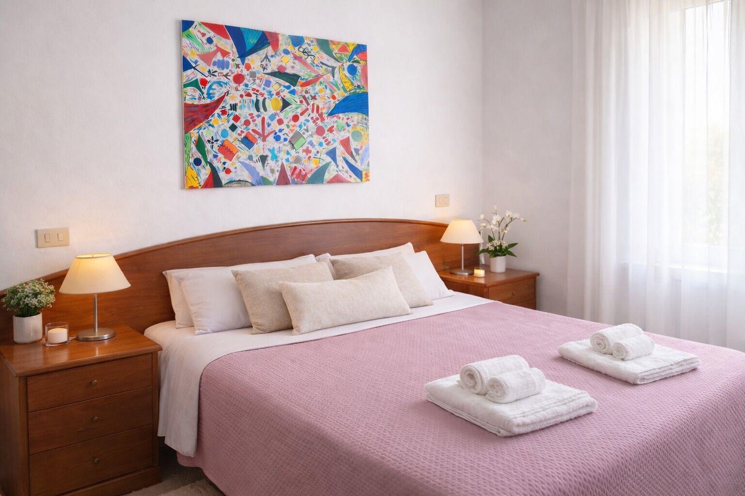 A hotel room with a wooden bed, a pink bedspread, white pillows, two bedside tables with lamps, a nightstand with a plant, and a colorful abstract painting on the wall.