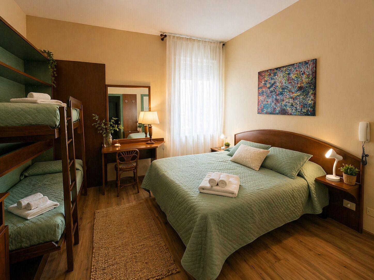 A hotel room with bunk beds, a desk, a chair, a bedside table with a lamp, a bedside plant, and a painting on the wall.