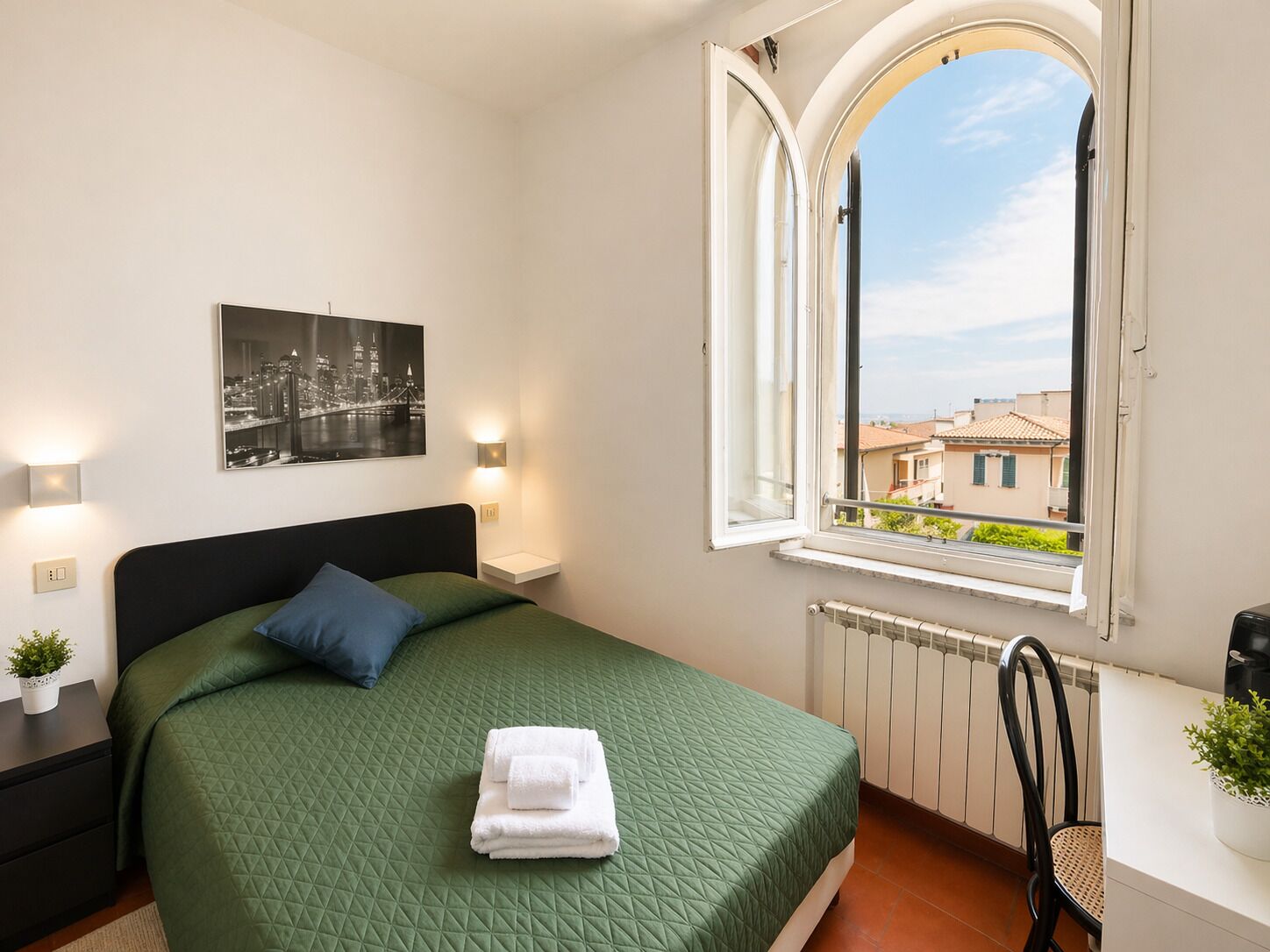Une chambre à coucher comprenant un lit, une table de chevet, une lampe fixée au mur, une fenêtre avec vue et un radiateur.