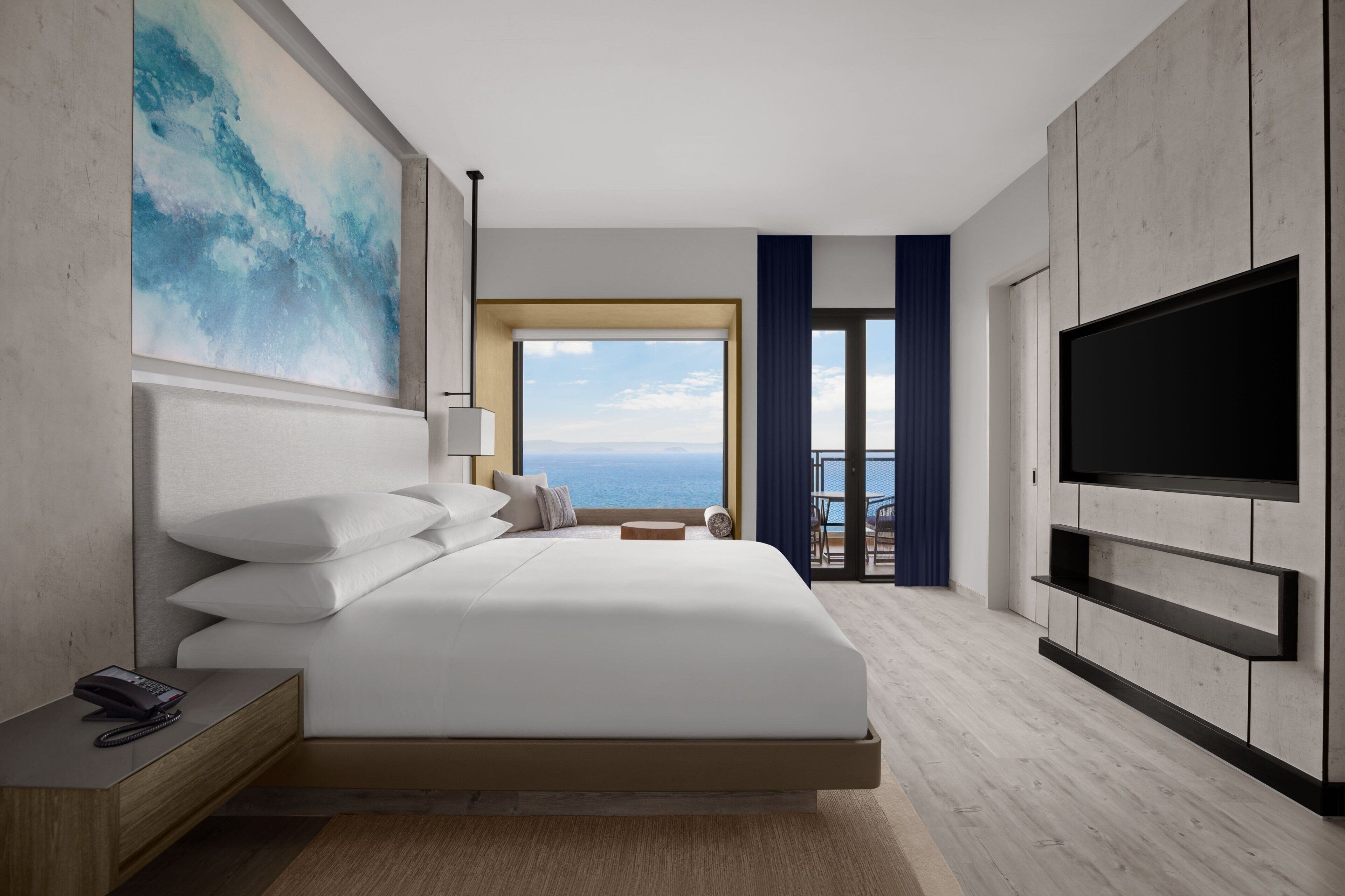 Habitación de hotel moderna con una cama grande, un televisor de pantalla plana, un cuadro en la pared y vista al océano a través de un ventanal amplio.
