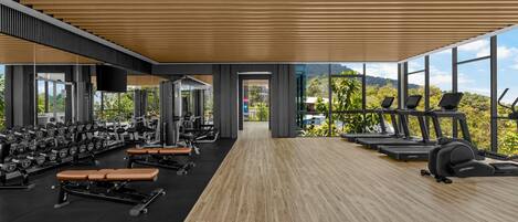 A modern gym with wooden flooring, exercise equipment, and large windows offering a view of greenery.