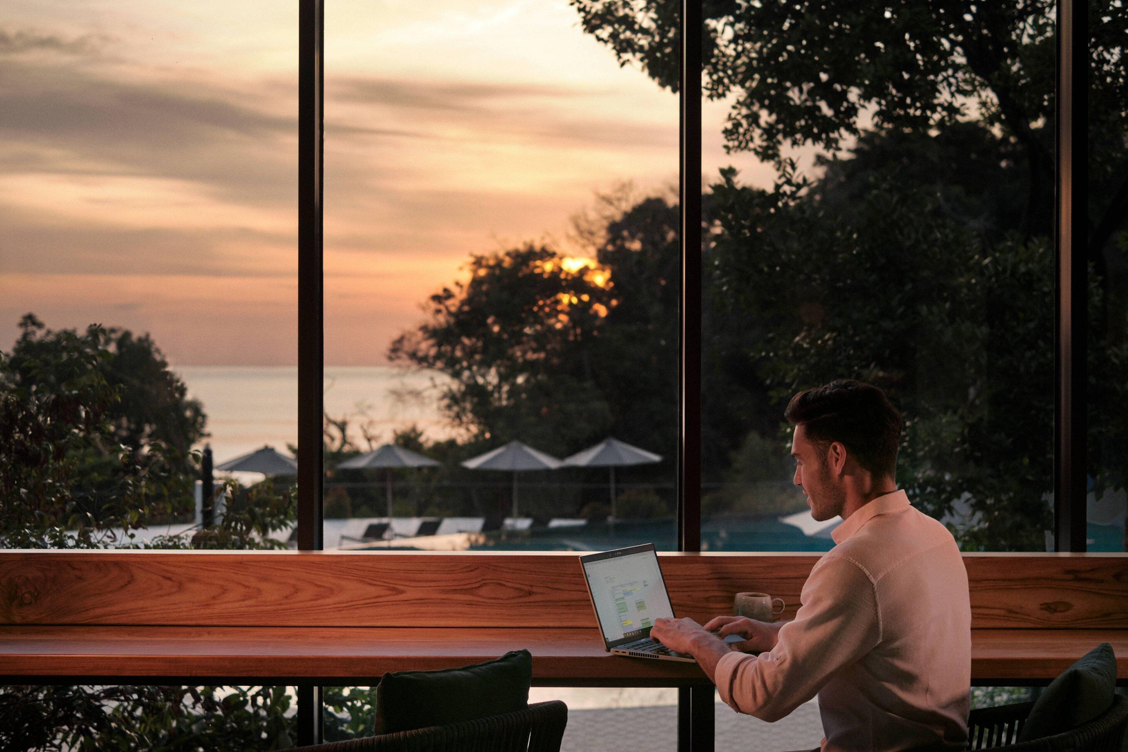 A man working on a laptop at a wooden table with a view of the sunset over the ocean.