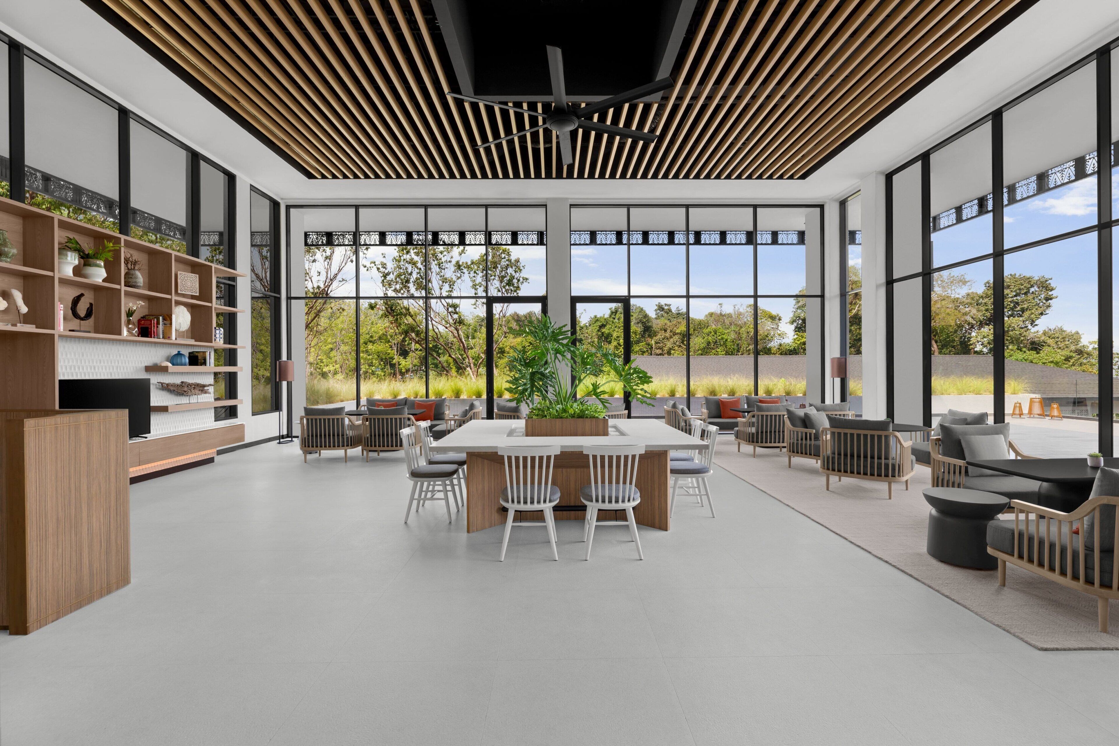A spacious, modern dining area with a central table surrounded by chairs, large windows offering a view of the outdoors, and a ceiling with a slatted design.