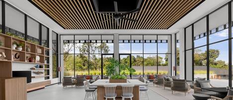 A spacious, modern dining area with a central table surrounded by chairs, large windows offering a view of the outdoors, and a ceiling with a slatted design.