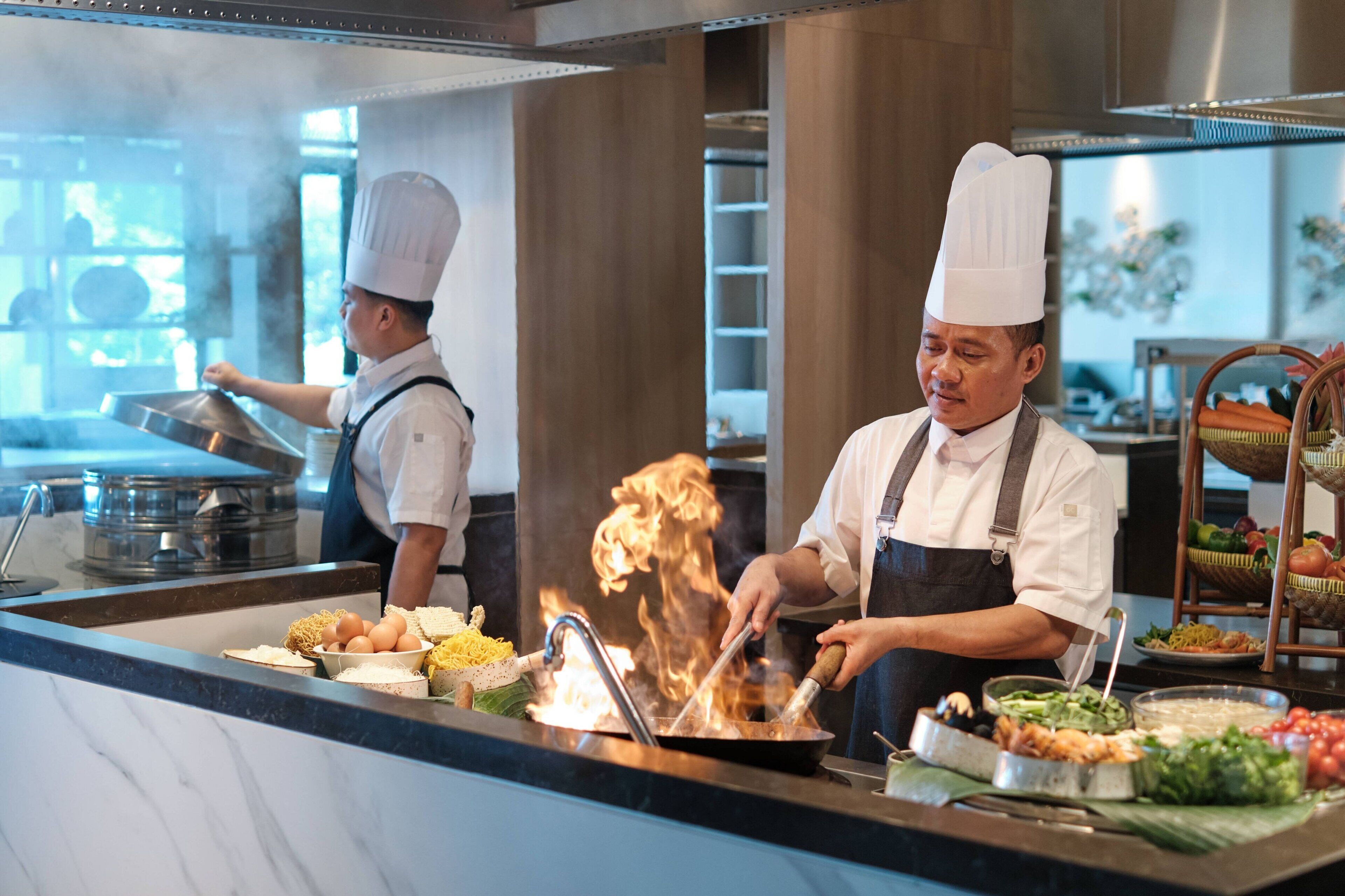 Chefs cooking in a professional kitchen with a variety of ingredients and cooking utensils visible.